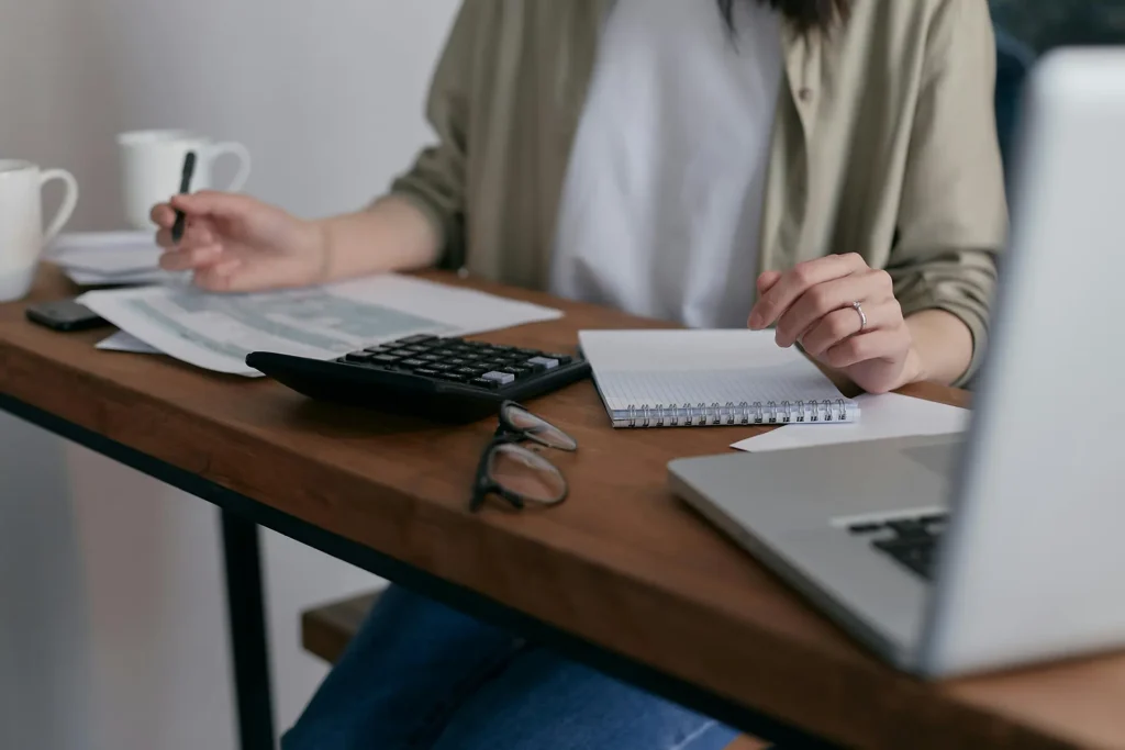 Person working at a wooden desk with calculator, notebook, glasses, and laptop while reviewing paperwork