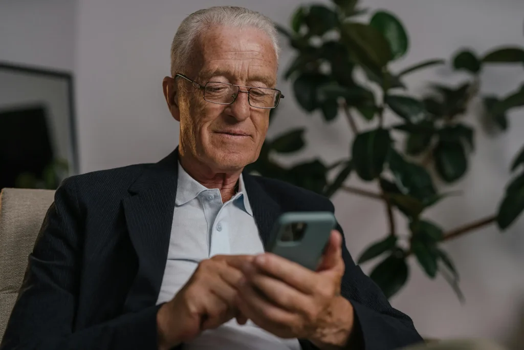 Senior man in a blazer using a smartphone while seated indoors, reviewing messages or pension details